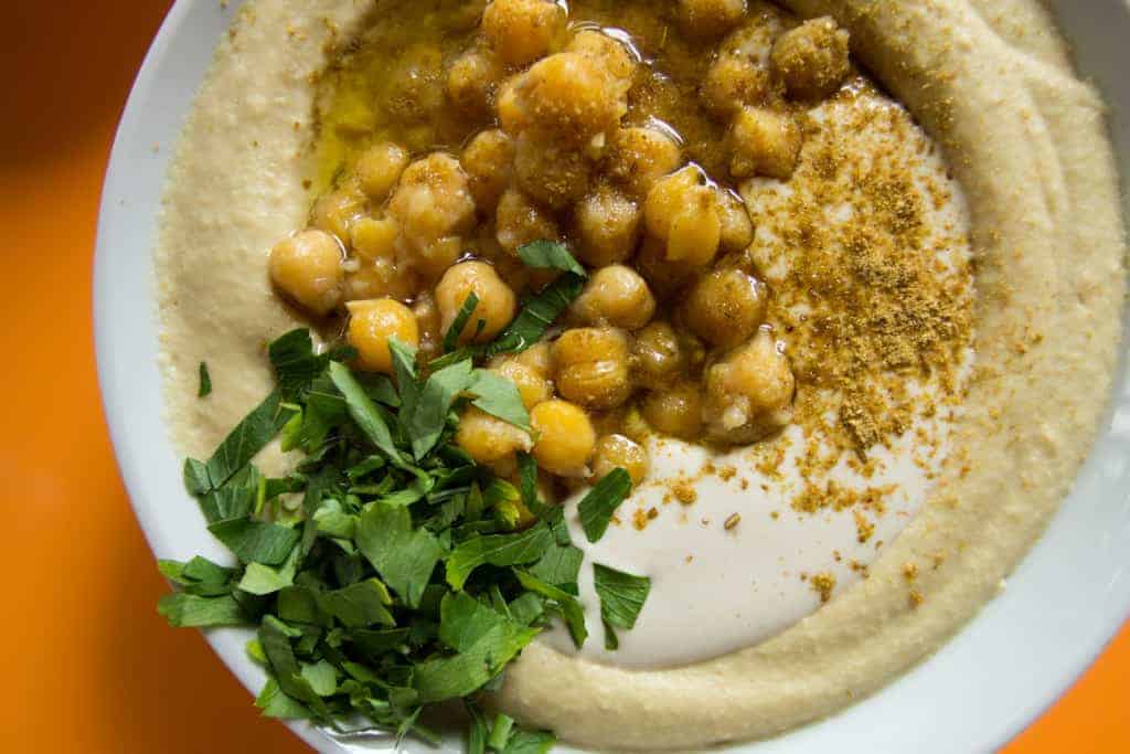 A bowl of hummus topped with chickpeas, herbs, olive oil, and spices on an orange background.