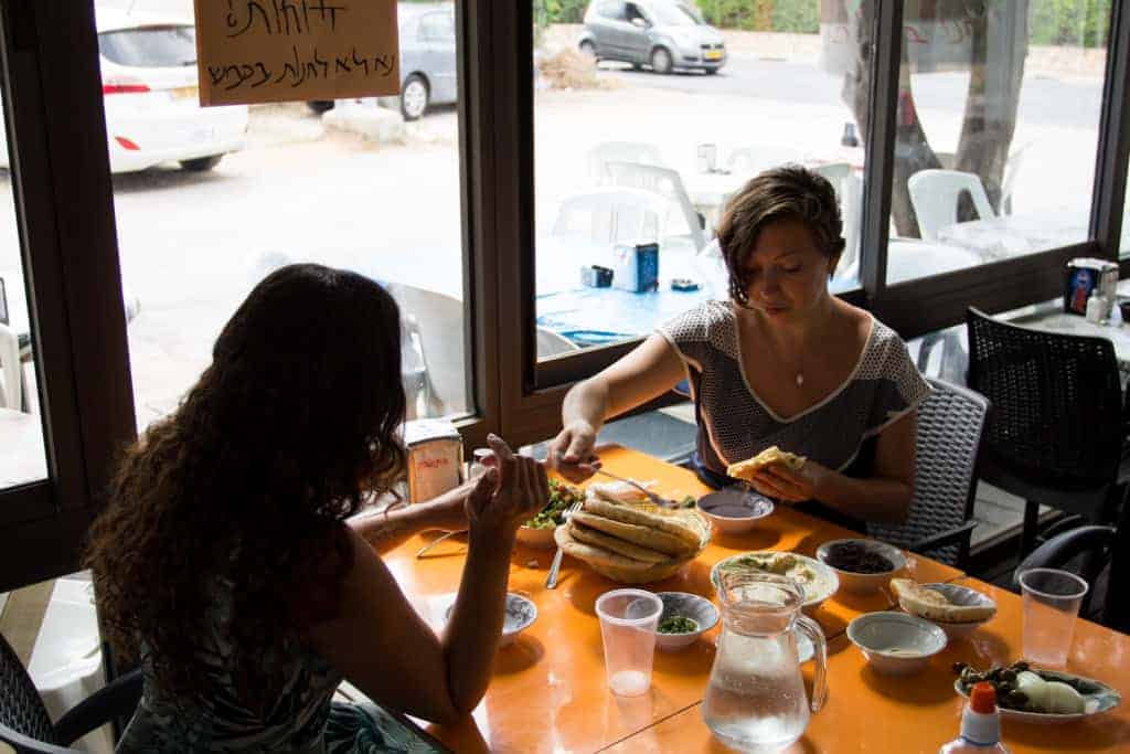 Two women sit at an orange table in a cafe, sharing a meal with bread, salads, and water by a large window.