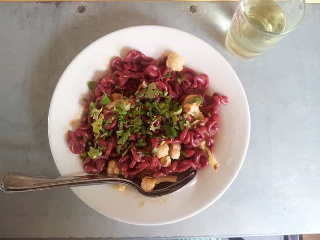 White plate of beet pasta with cauliflower and herbs, fork and spoon on the side, next to a glass of white wine.