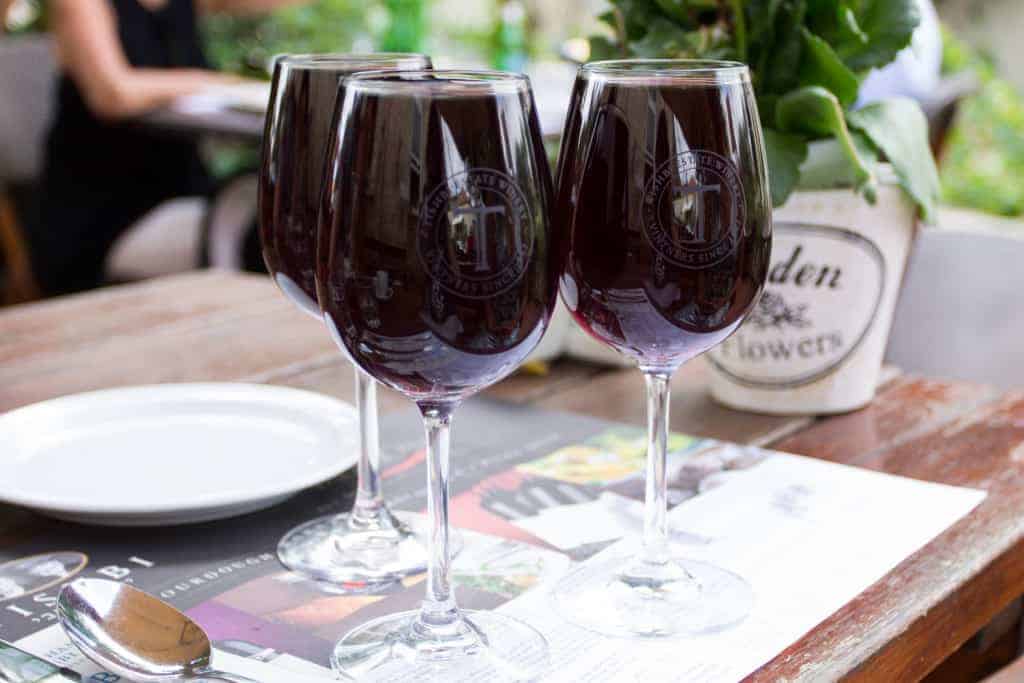 Three glasses of red wine on a wooden table with a plate, spoon, and potted plant nearby.