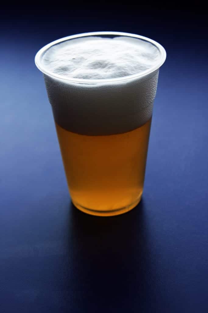 A plastic cup filled with golden beer topped with a foamy head, set against a dark blue background.