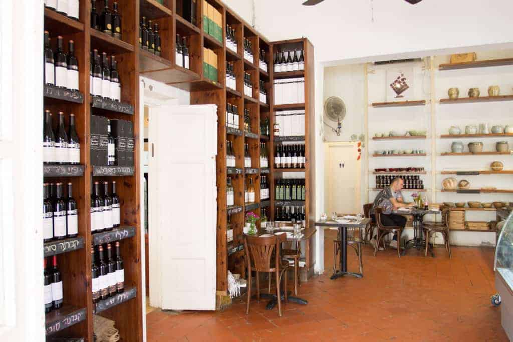 Wooden shelves filled with wine bottles in a cozy café; one person sits at a table near the back.