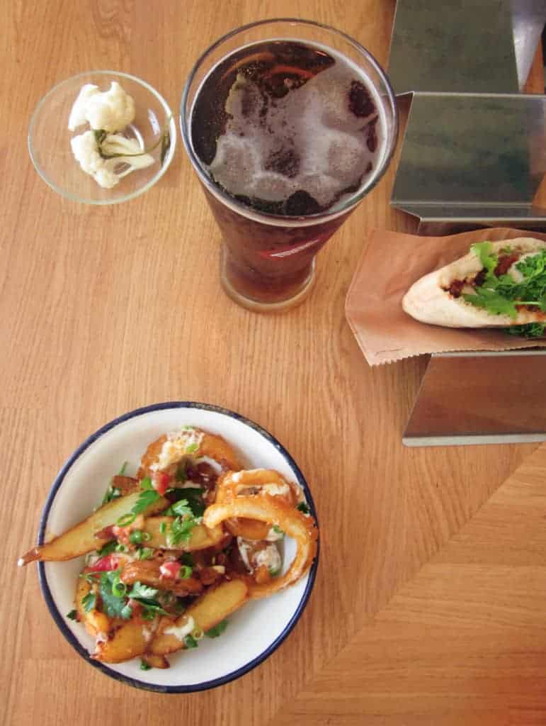 A plate of loaded fries, a hot dog in a stand, a glass of soda, and a small bowl of garlic on a wooden table.