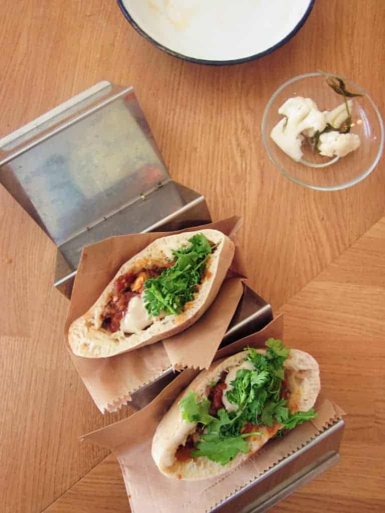 Two pita sandwiches with greens and sauce on a wooden table, next to a small dish of pickled garlic.