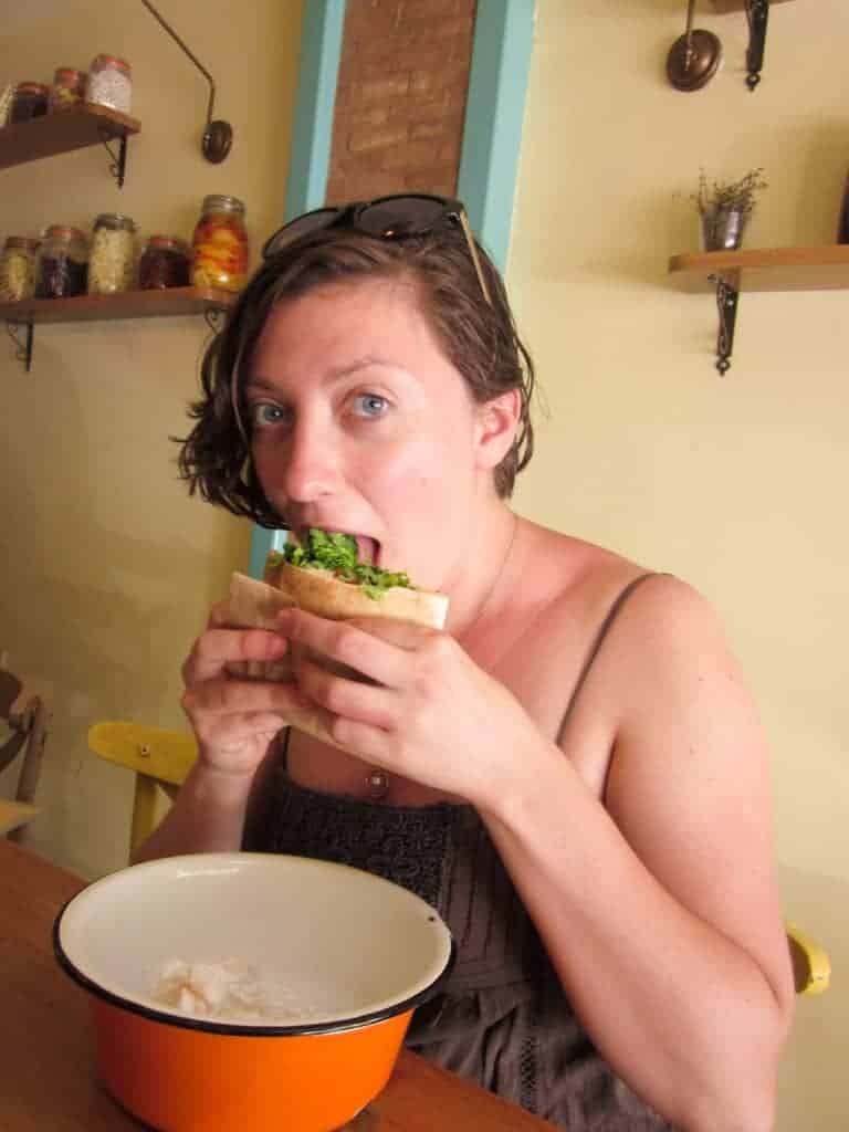 Woman with short hair eating a sandwich with greens, sitting at a table in a cozy, rustic cafe.