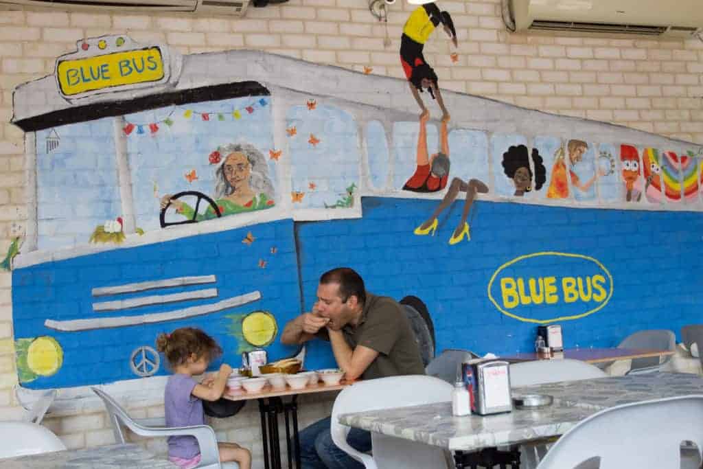 A man and a young girl eat at a table in front of a mural of a blue bus with cartoon people.