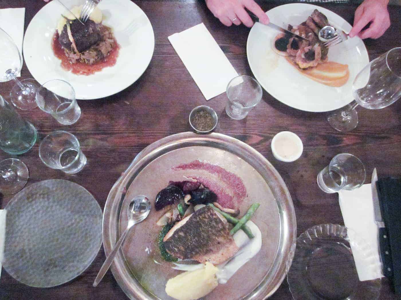 Three plates of food on a wooden table, with two people eating and several empty glasses around.