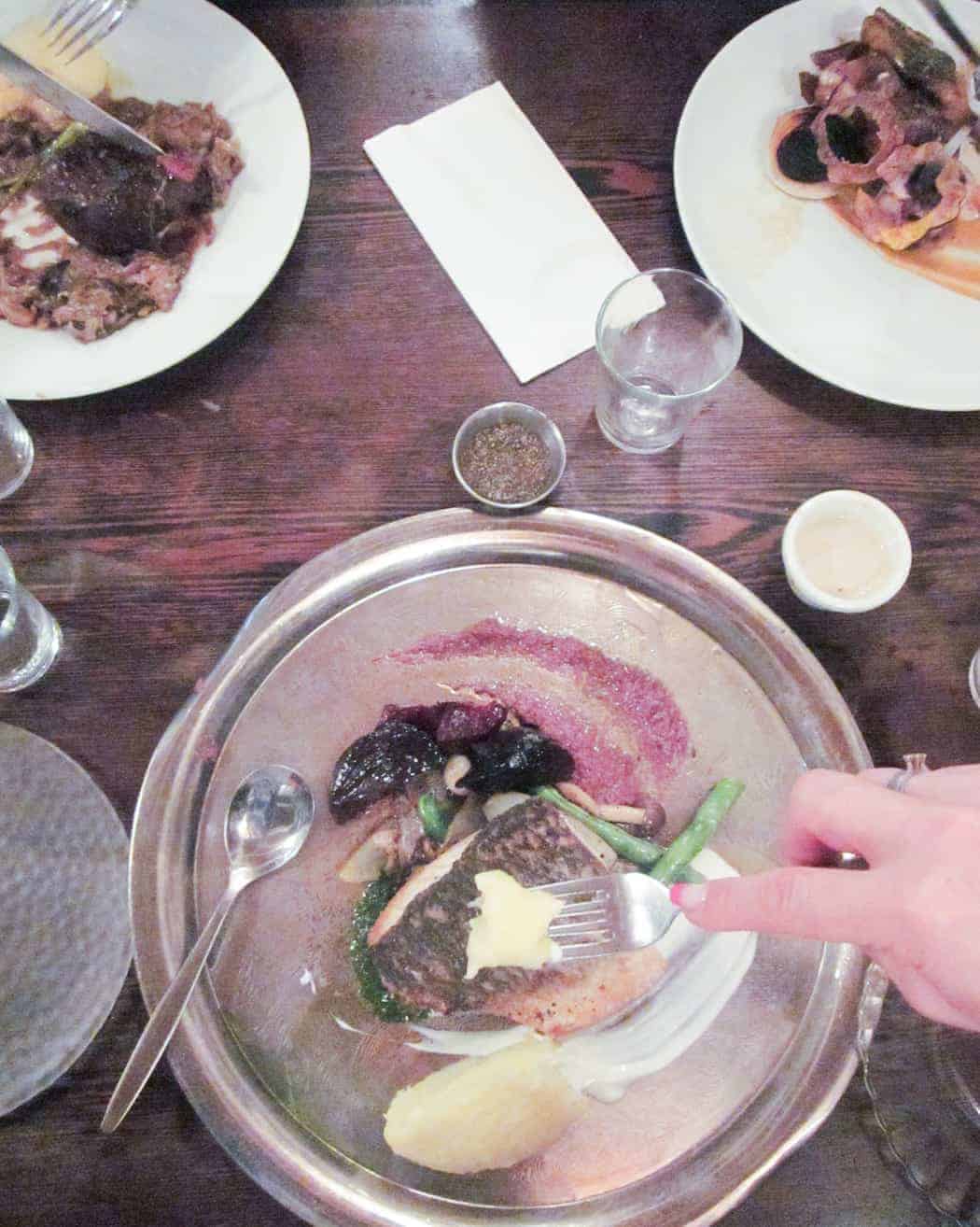 A person cutting fish with vegetables and mashed potatoes on a plate, surrounded by other dishes on a wooden table.