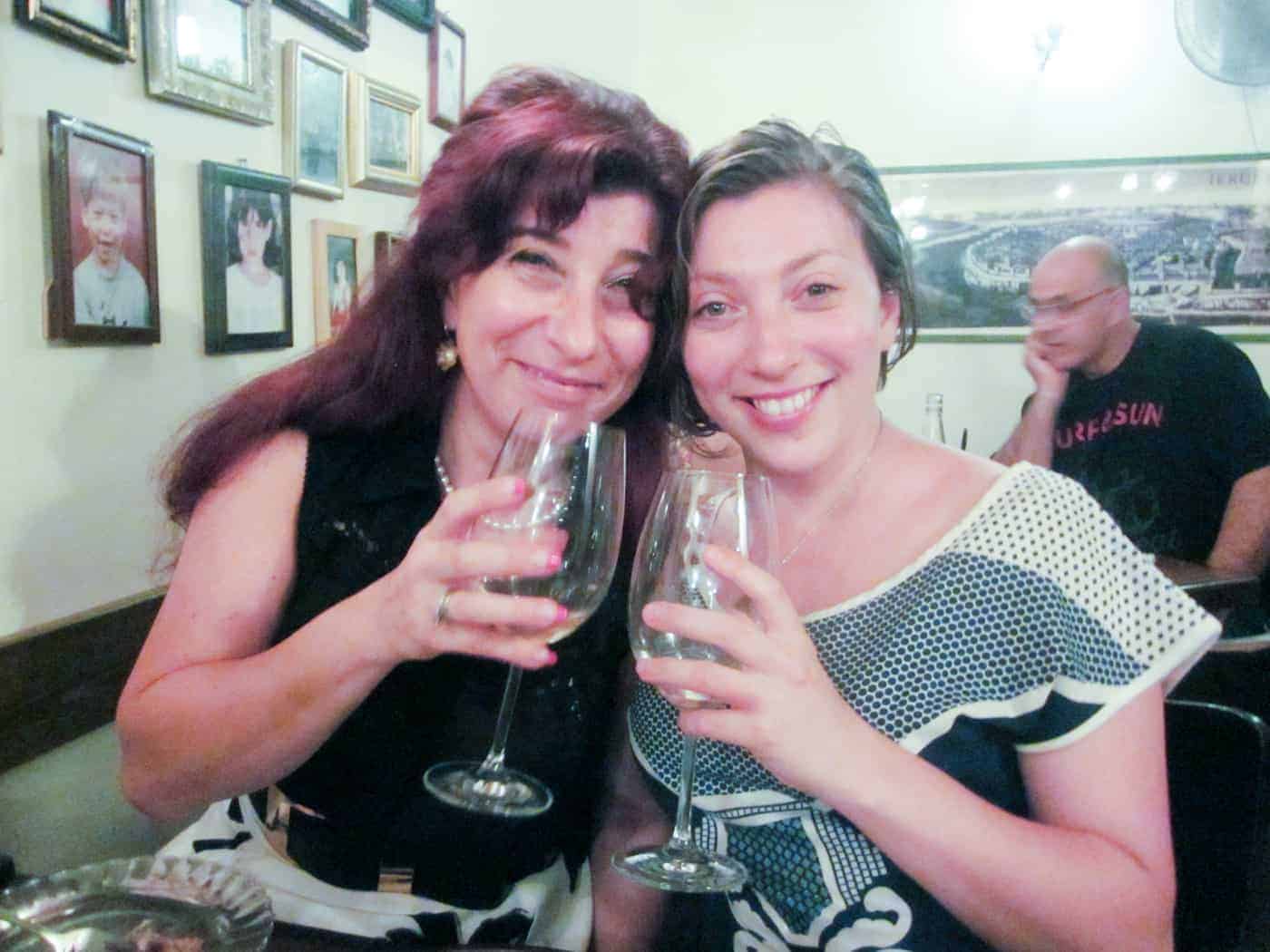 Two women smiling and holding wine glasses in a cozy, framed-photo-filled restaurant.