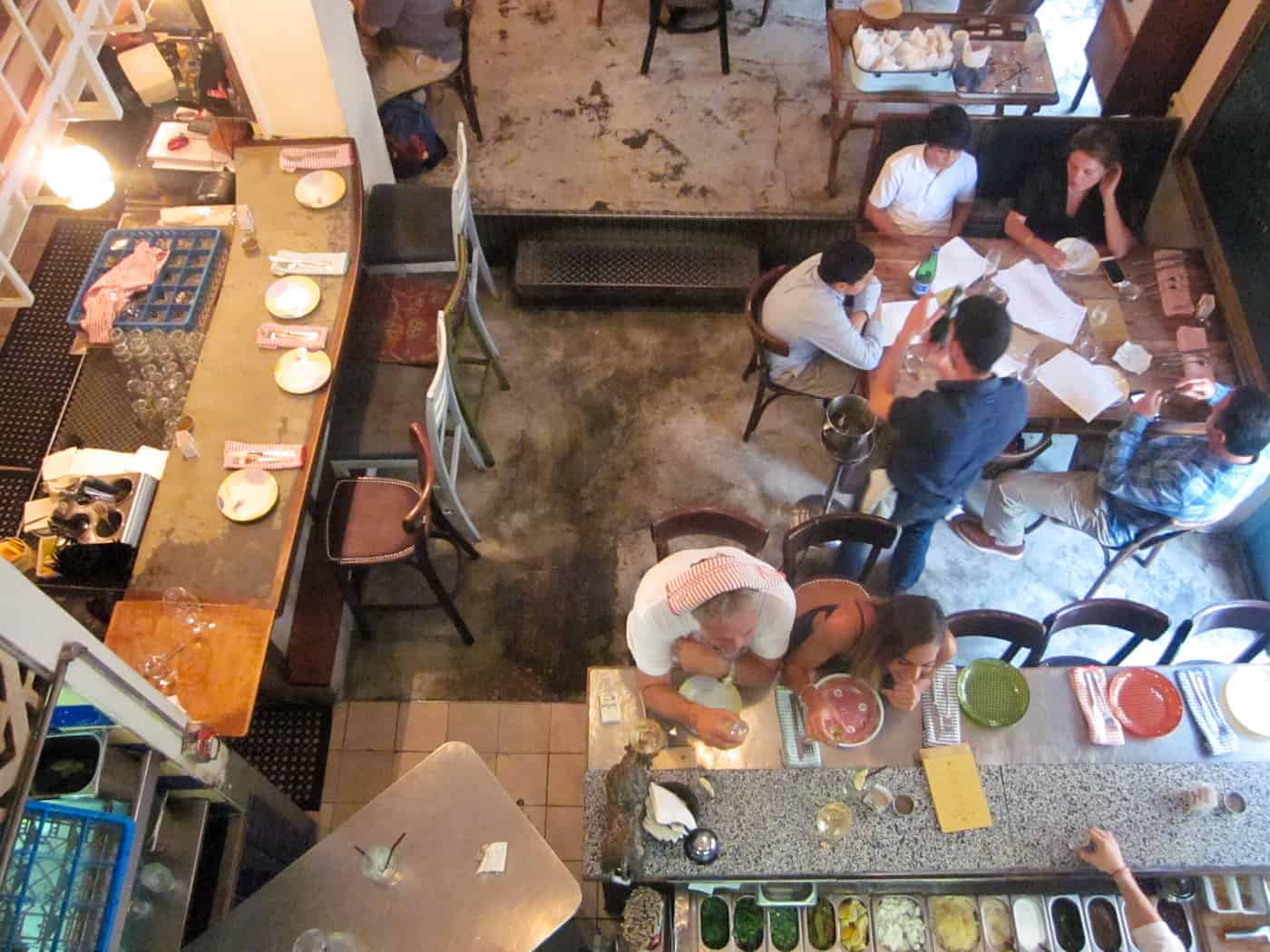 Overhead view of people dining and talking at tables in a casual restaurant with a counter and food prep area.