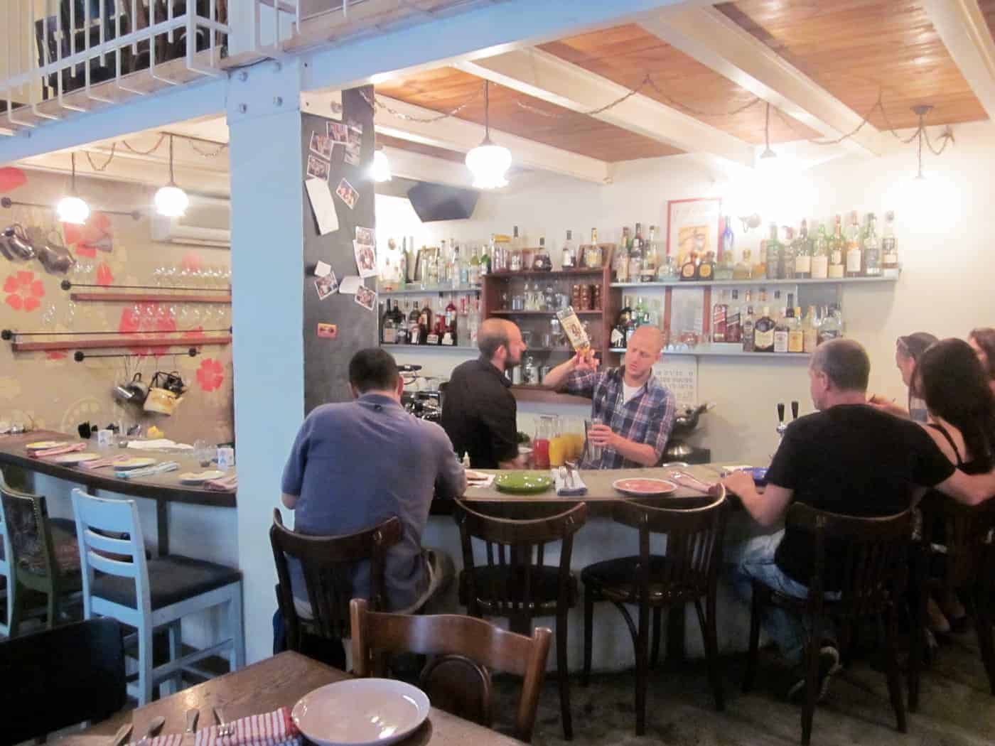 Bartender serving drinks to people seated at a cozy bar with shelves of bottles and warm lighting.