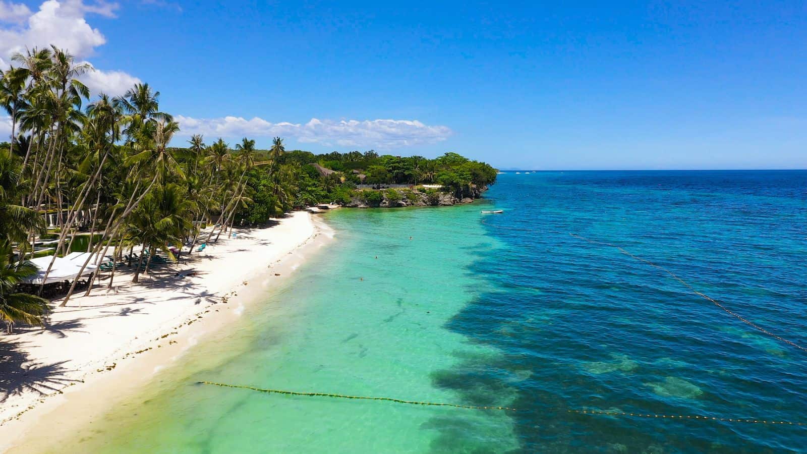 A sandy beach lined with palm trees borders clear turquoise water, with the shoreline curving gently toward a lush, green area under a bright blue sky.