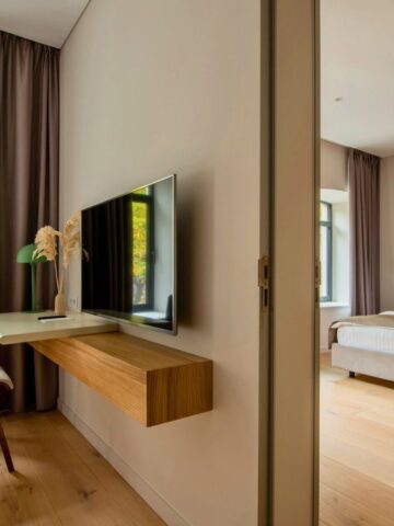 Modern hotel room with a wall-mounted TV, desk, chair, large bed, wooden floor, and bright windows.