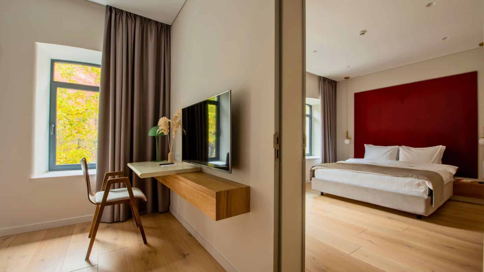 Modern hotel room with a wall-mounted TV, desk, chair, large bed, wooden floor, and bright windows.