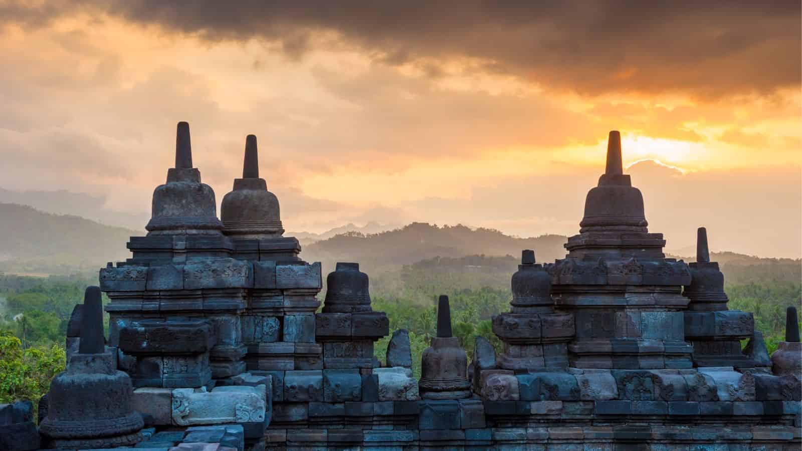 Ancient stone stupas at Borobudur Temple silhouetted against a vibrant sunrise sky with distant hills.