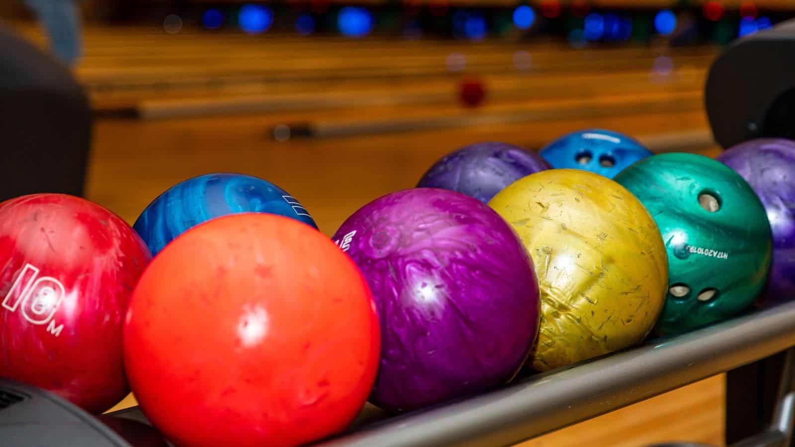 A row of colorful bowling balls, reminiscent of confiscated items at airport security, sits on a rack with bowling lanes and pins blurred in the background.