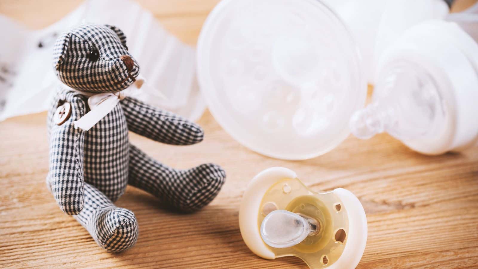 A checkered teddy bear, baby pacifier, and bottle parts on a wooden surface.