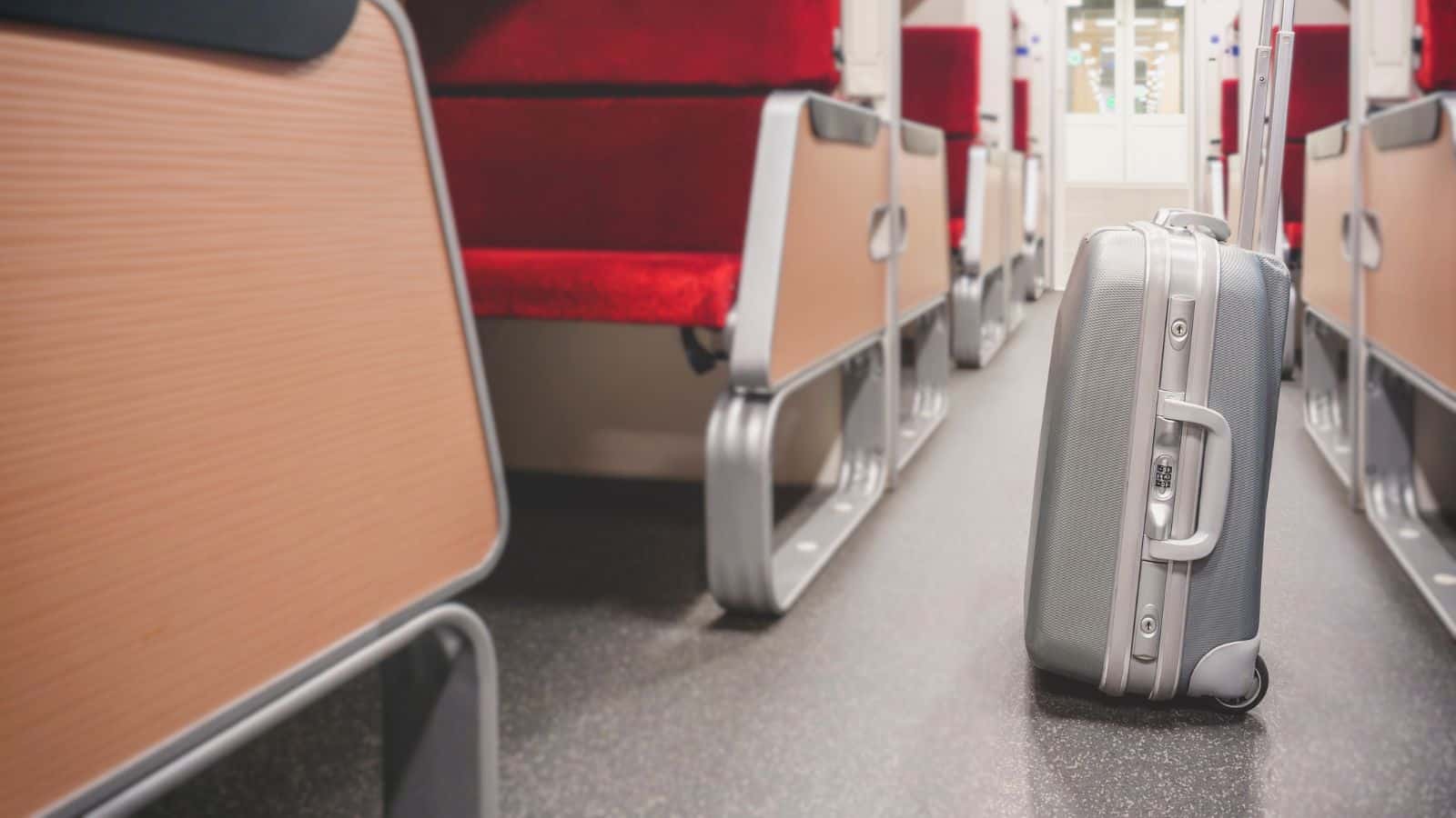 A silver hard-shell suitcase stands upright in the aisle between rows of empty red seats inside a train carriage—a gentle reminder to follow train etiquette for a smoother train ride.