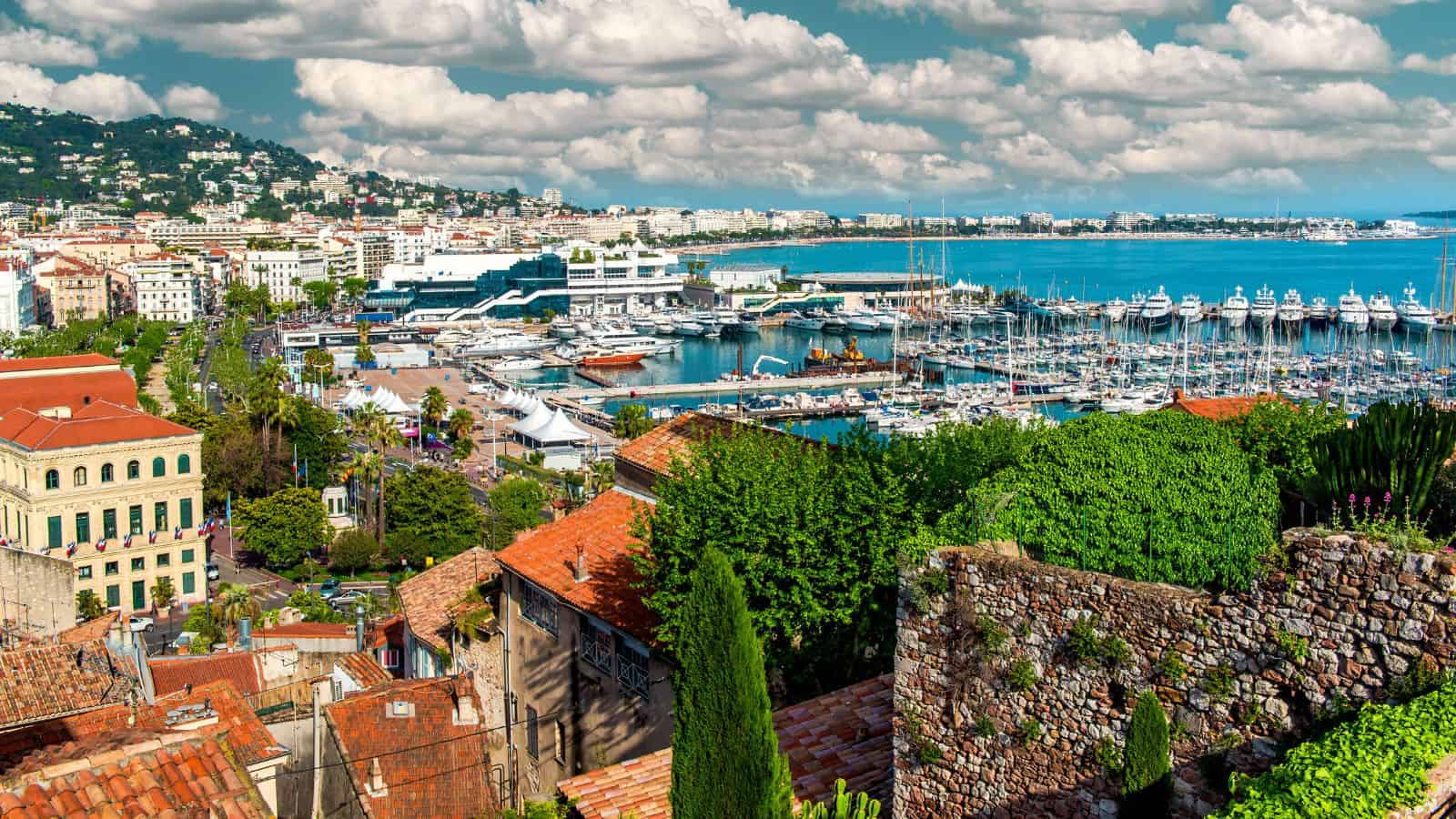 Scenic view of Cannes harbor with yachts, old town rooftops, greenery, and the sea under a partly cloudy sky.