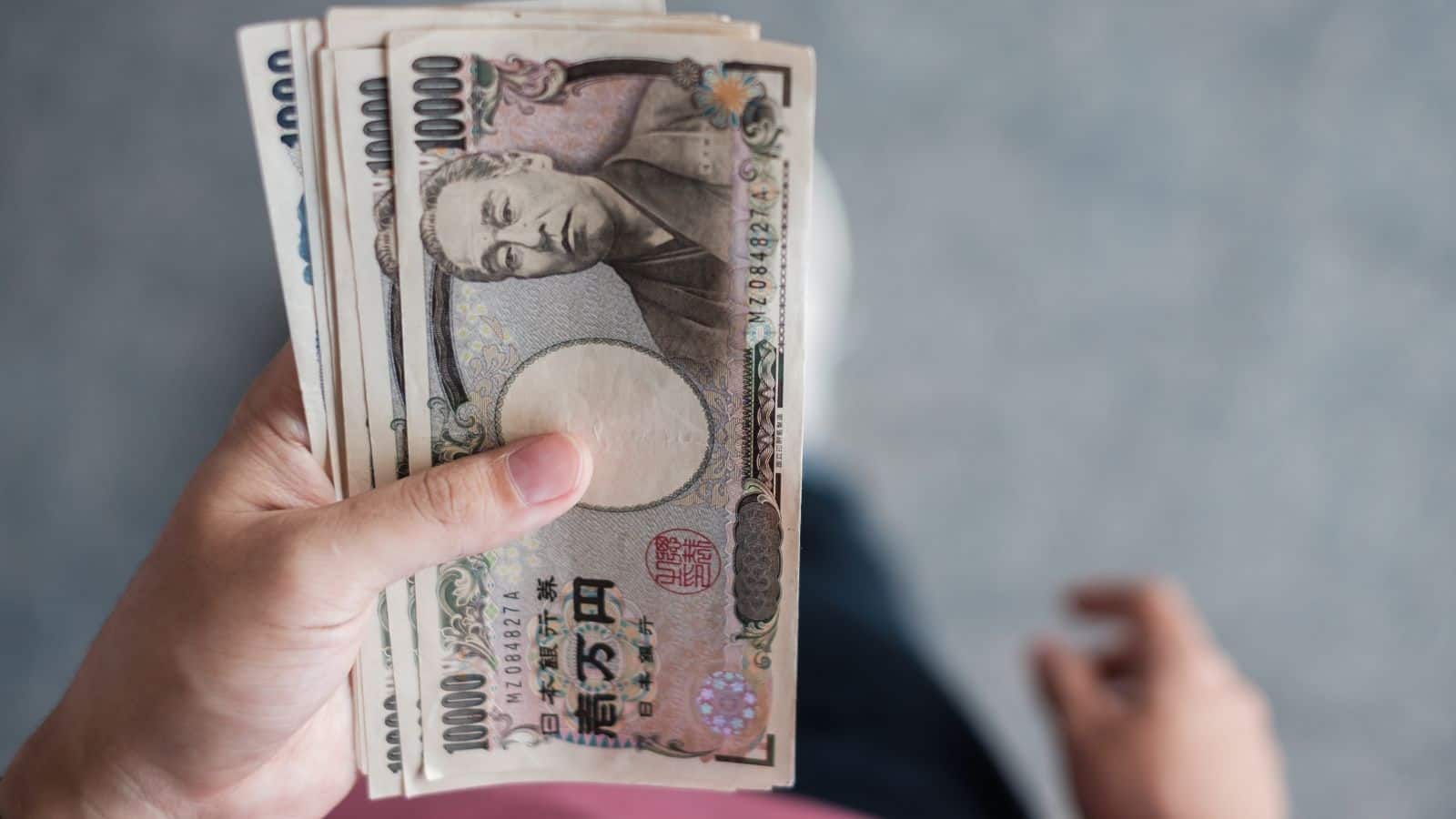 A hand holds several Japanese yen banknotes, viewed from above against a blurred background.