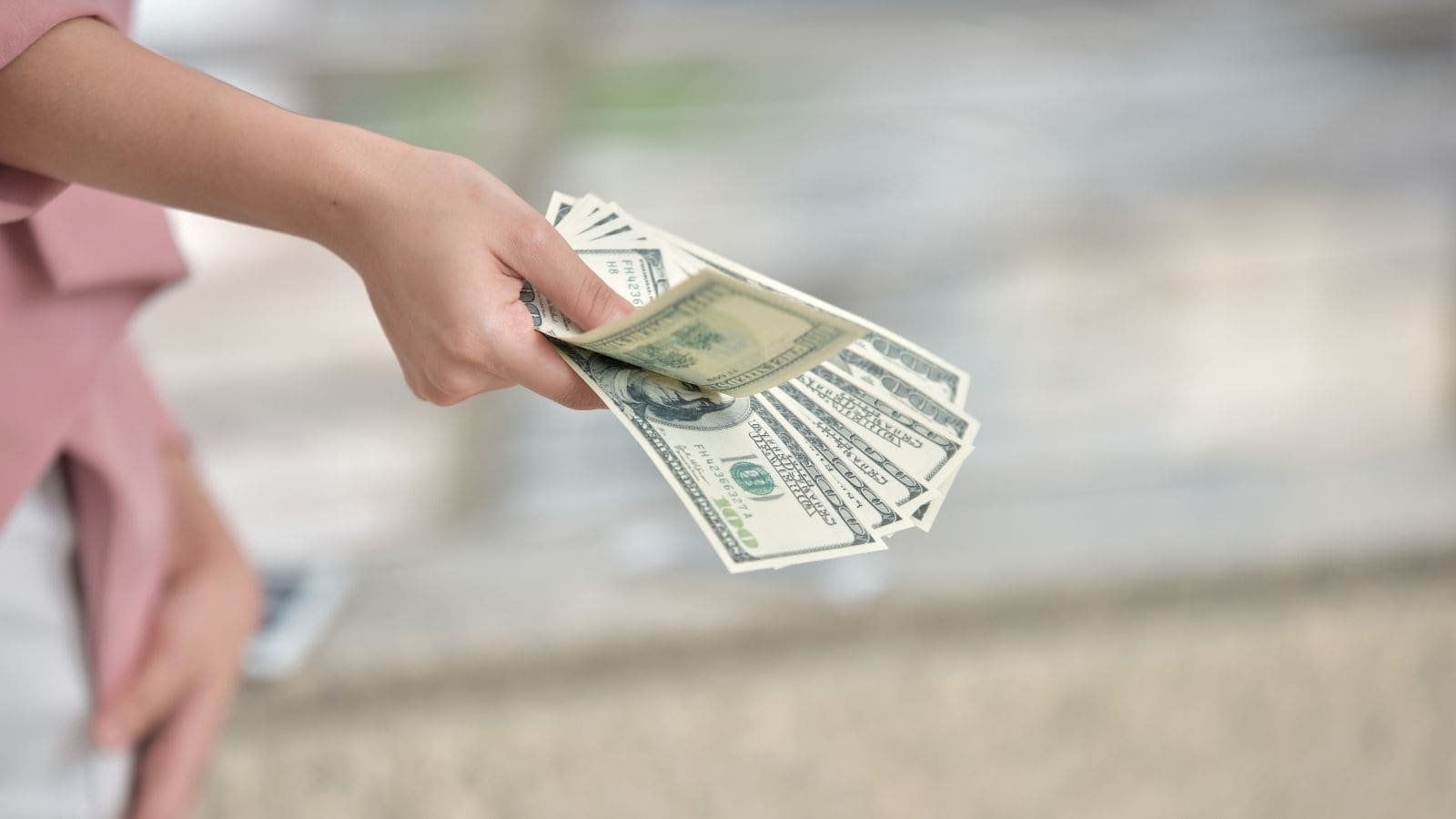 A person’s hand holding several spread-out U.S. hundred-dollar bills, reflecting the travel confusion first-time travelers may feel when adjusting from European habits, with a blurred background.