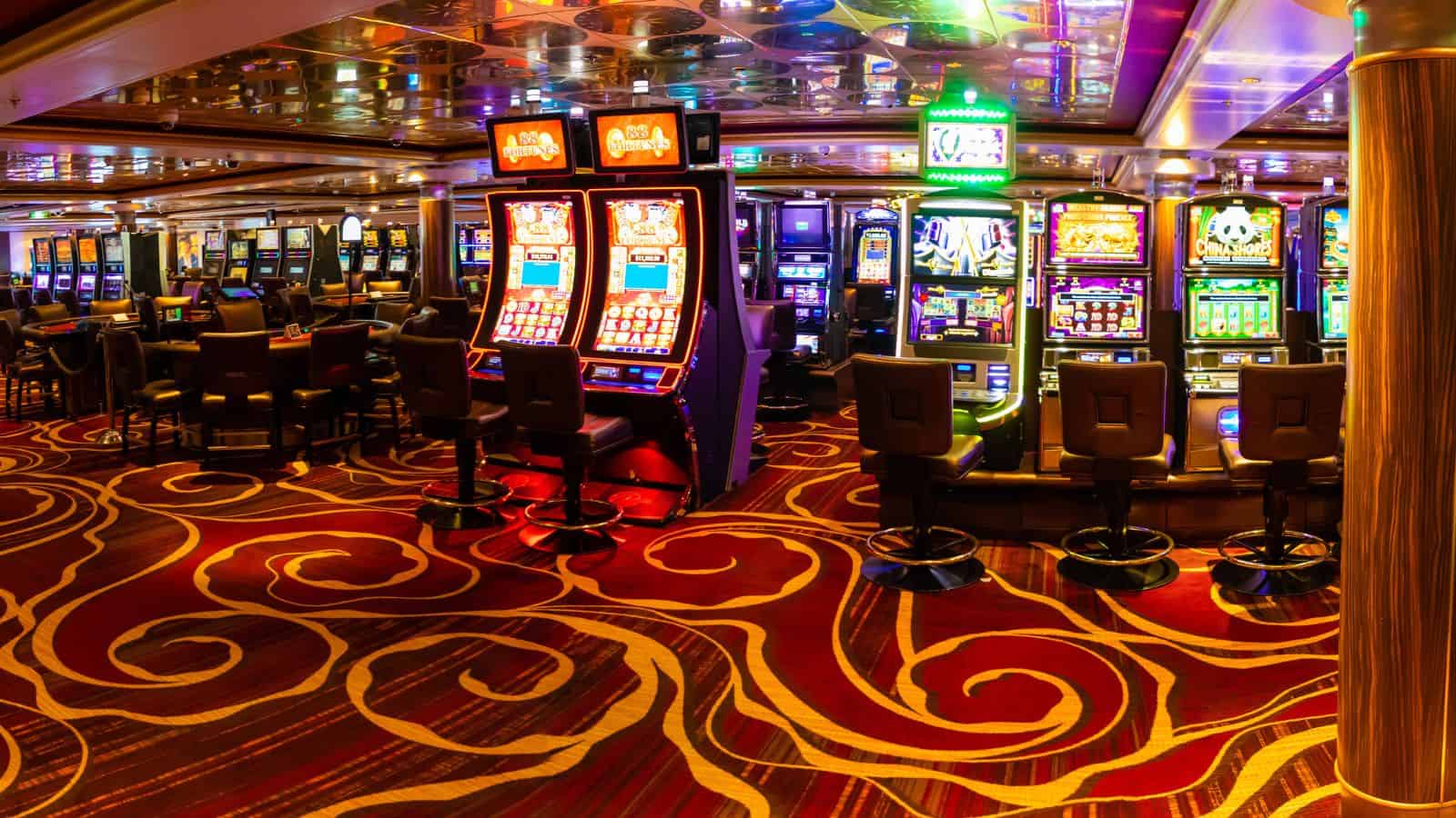 A row of slot machines in a brightly lit casino with patterned carpet and empty chairs.
