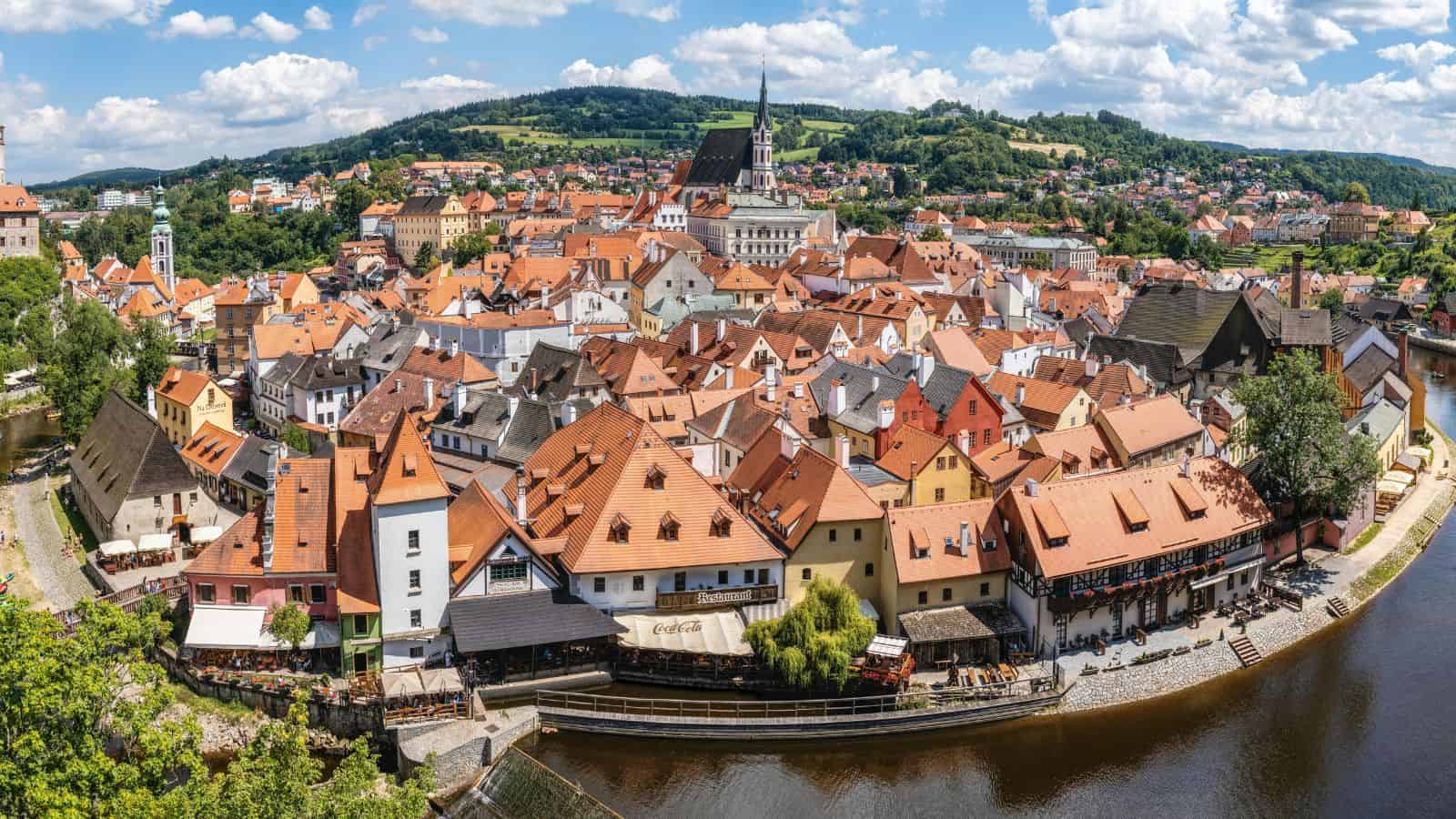 Panoramic view of a European town with red-roofed buildings by a river, surrounded by green hills.