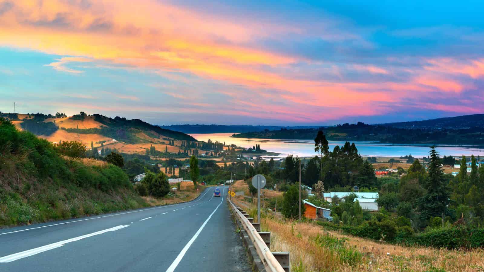 A scenic road leads to a lake at sunset, with colorful clouds and hills in the background.