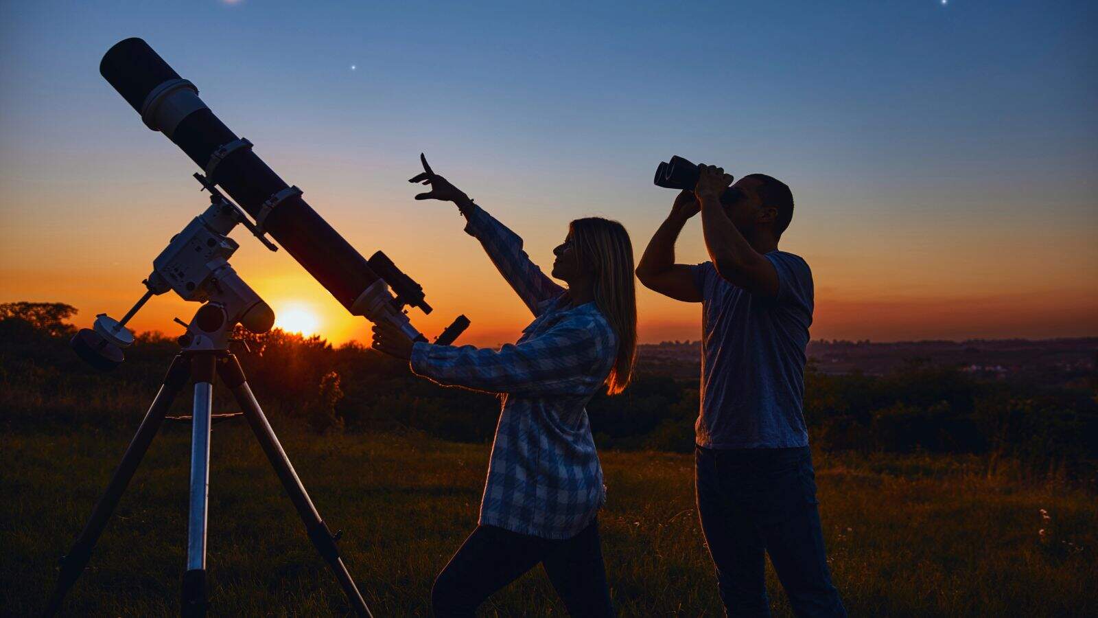 Two people stand outdoors at sunset, one looking through a telescope and the other pointing at the sky while holding a notebook, with grassy fields in the background.