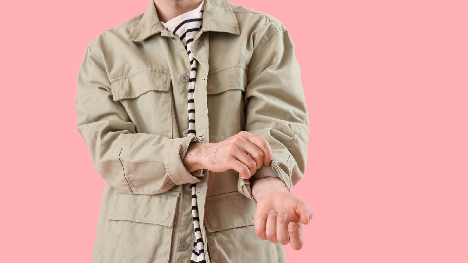 A person wearing a beige jacket over a striped shirt is rolling up their sleeve against a plain pink background, as if searching for commonly lost items often left behind at U.S. airports. Their face is partially out of frame.