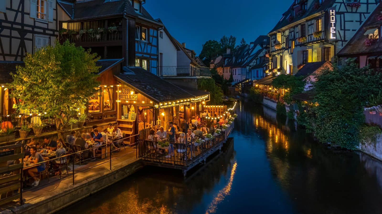 People dining at a riverside restaurant with warm lights in a charming European town at dusk.