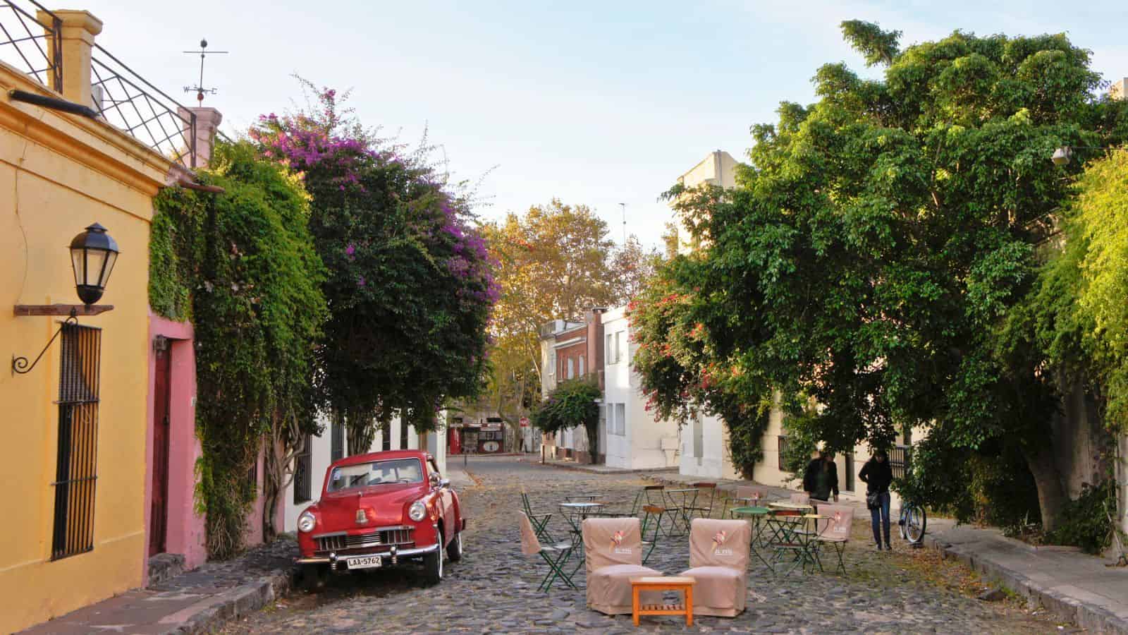 Cobblestone street with a red vintage car, outdoor café tables, and colorful buildings lined with greenery.