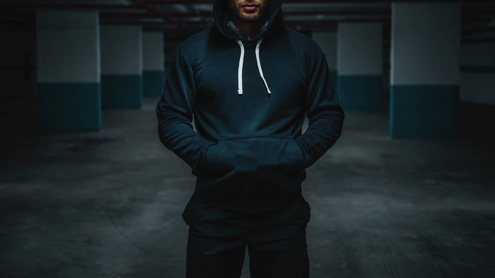 Person in a dark hoodie stands with hands in pockets in a dimly lit, empty parking garage.