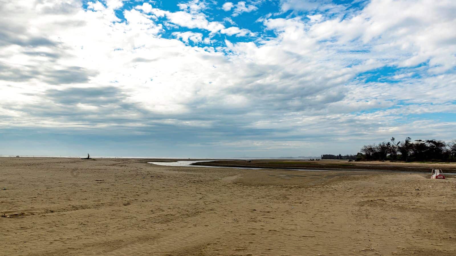 A sandy beach with a winding stream flowing toward the sea under a partly cloudy sky. Some trees are visible in the distance on the right side.