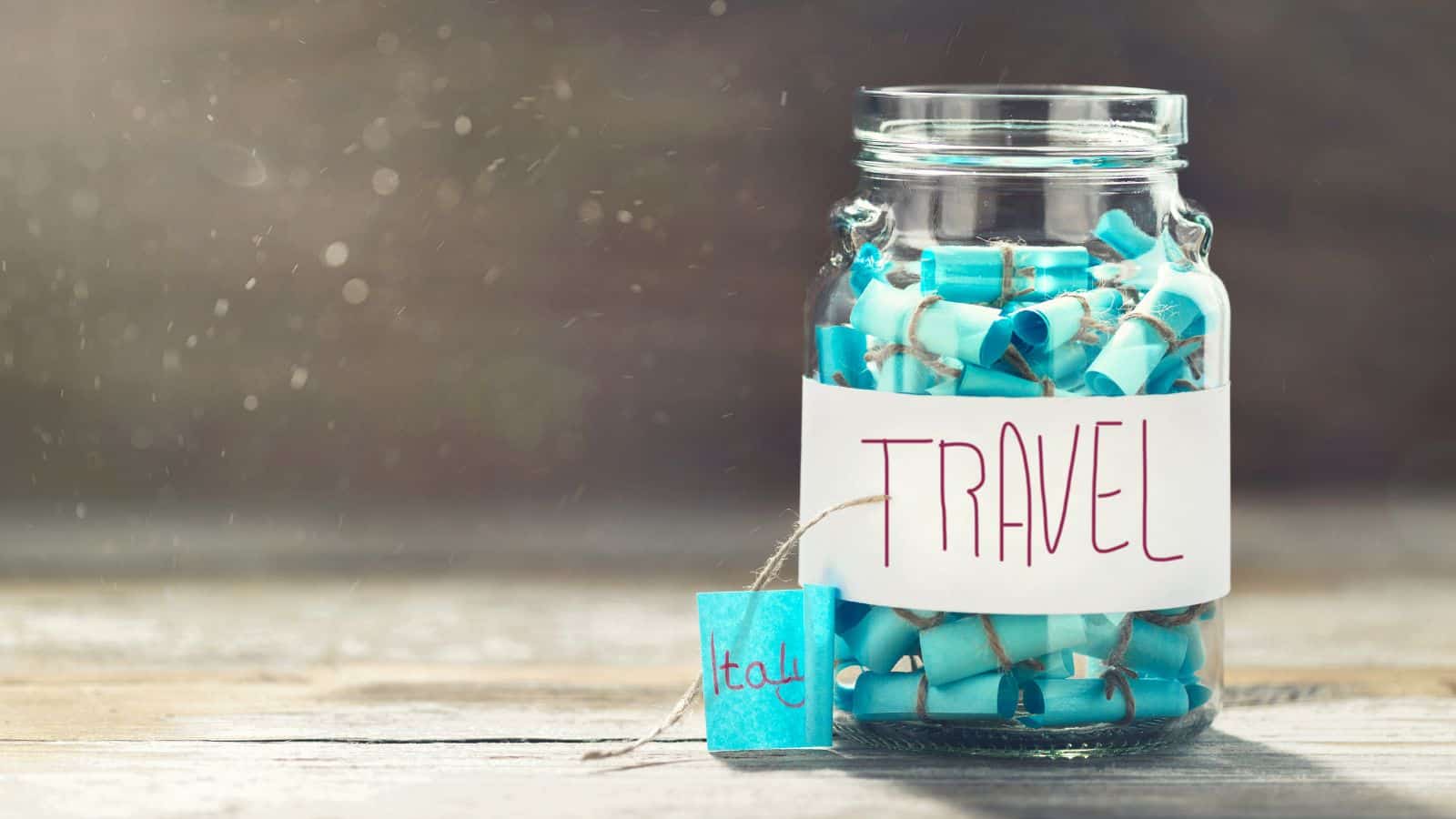 A glass jar labeled "TRAVEL" filled with rolled-up blue and teal paper scrolls sits on a wooden surface. A tag reading "Italy" is attached to the jar. Sunlight streams in, creating a soft, bright atmosphere.