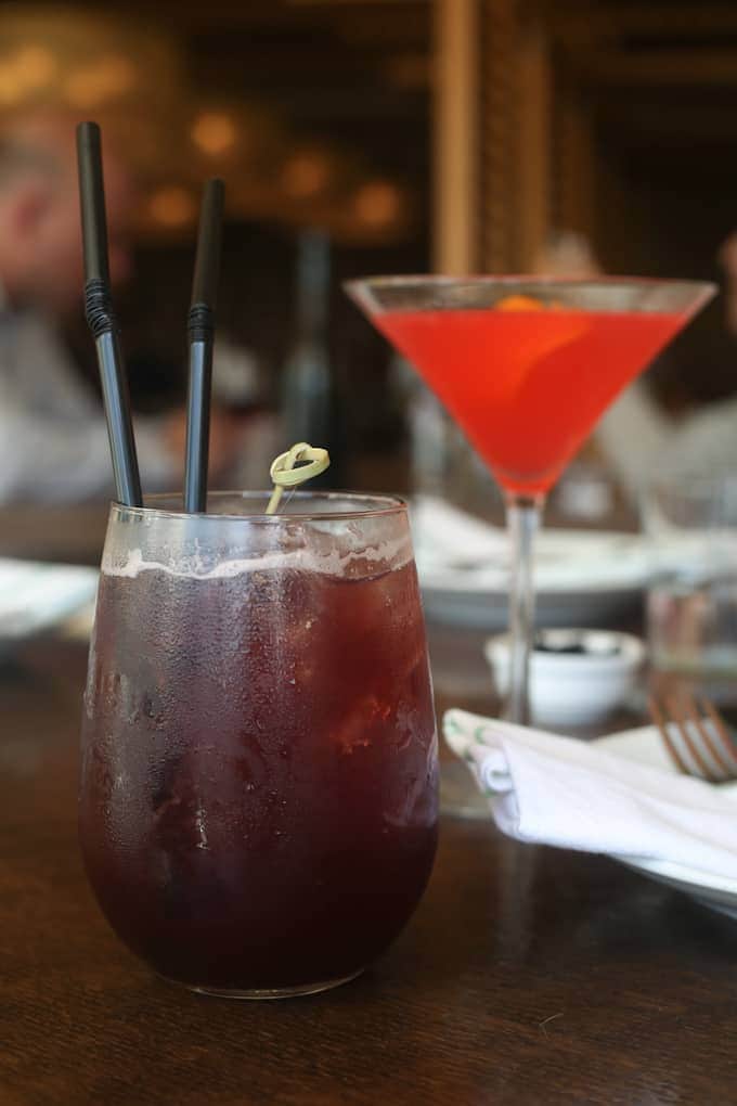 Two colorful cocktails on a wooden table, with blurred people and restaurant background.