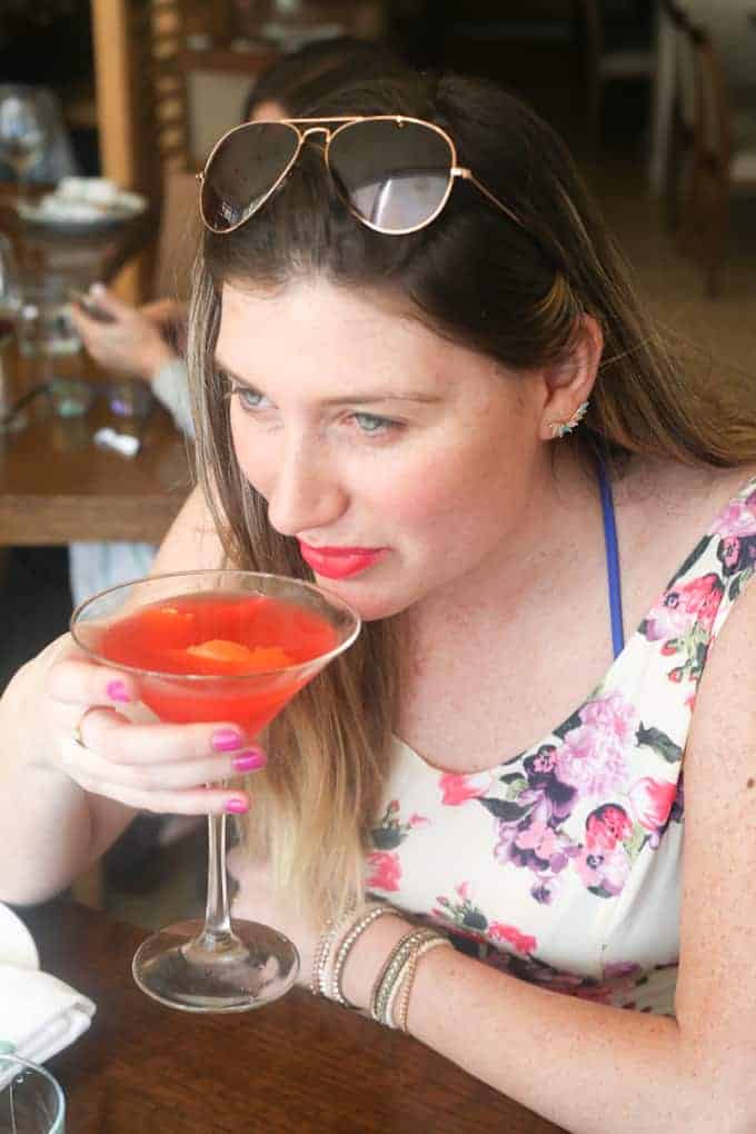 Woman with sunglasses on her head sips a red cocktail at a restaurant, wearing a floral dress.