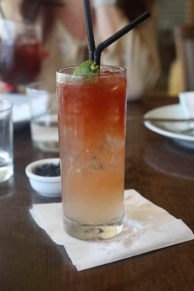 A tall iced drink with a lime wedge and two straws sits on a napkin on a wooden table.