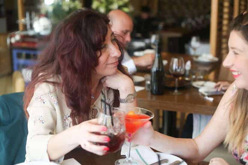 Two women sitting at a restaurant table, smiling and clinking glasses with colorful drinks.