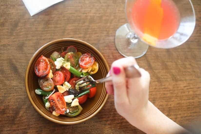 A hand with pink nail polish holds a fork over a bowl of tomato salad next to a pink cocktail.