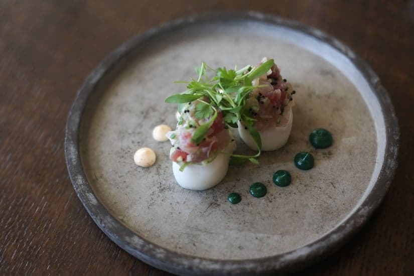 Elegant plated appetizer with two rounds topped with greens, dotted with green and beige sauces on a gray plate.