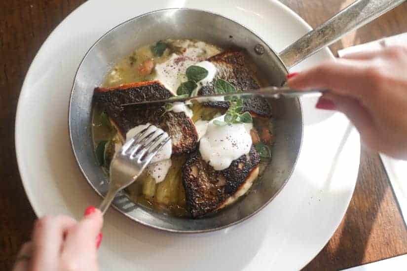 Person cutting cooked fish with crispy skin and sauce in a metal pan, garnished with herbs.