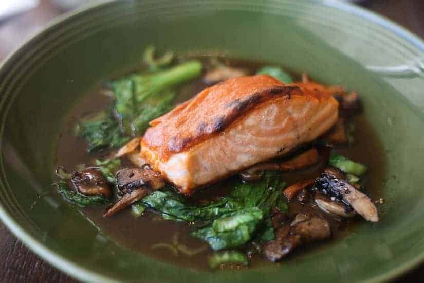 Grilled salmon fillet on a bed of greens and mushrooms in a brown sauce, served on a green plate.