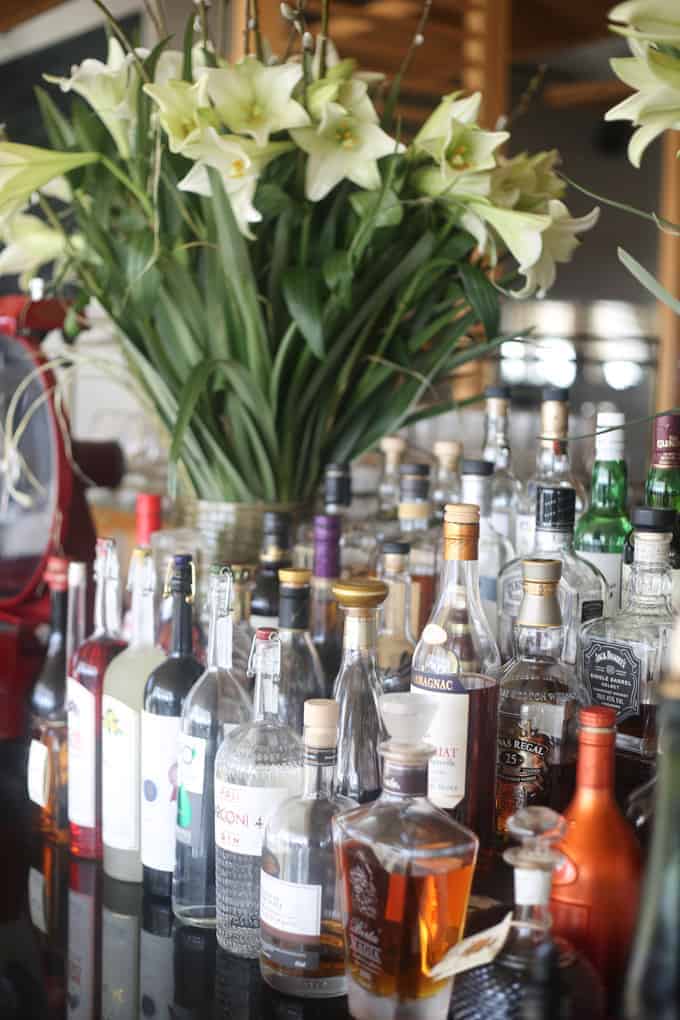 Assorted liquor bottles on a bar counter with a large vase of white flowers in the background.