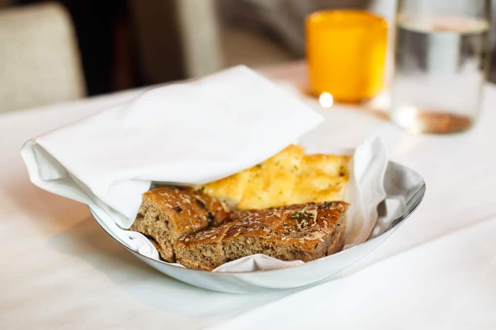 A bread basket with two types of bread, partially covered by a white napkin, sits on a table next to a glass of water and a yellow candle holder.