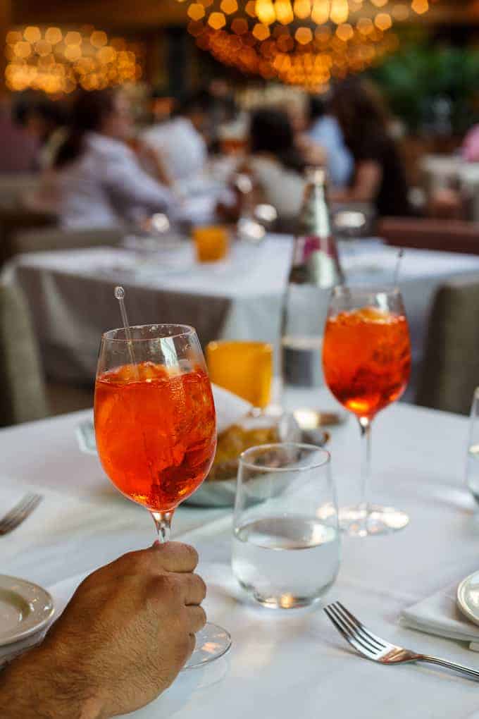 A hand holds a glass of orange cocktail on a white table set with another cocktail, a glass of water, and a bottle. Other tables and diners are visible in the blurred background.