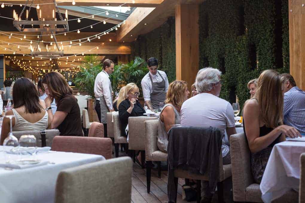 People are dining at a restaurant with string lights overhead, greenery on the walls, and wooden beams. A waiter in a gray apron stands by a table, while diners sit and talk at tables covered with white tablecloths.