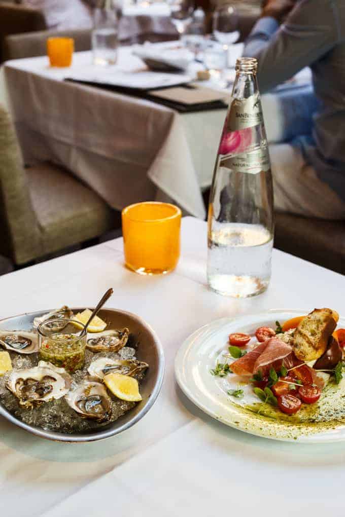 A restaurant table with a plate of oysters on ice with lemon wedges, a plate of prosciutto, bread, and cherry tomatoes, a bottle of sparkling water, an orange tumbler, and menus in the background.
