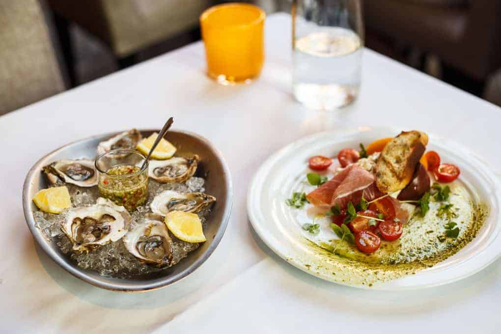 A plate of oysters on ice with lemon wedges and sauce sits next to a plate of prosciutto, cherry tomatoes, mozzarella, greens, and toasted bread on a white tablecloth. An orange glass and a water bottle are in the background.