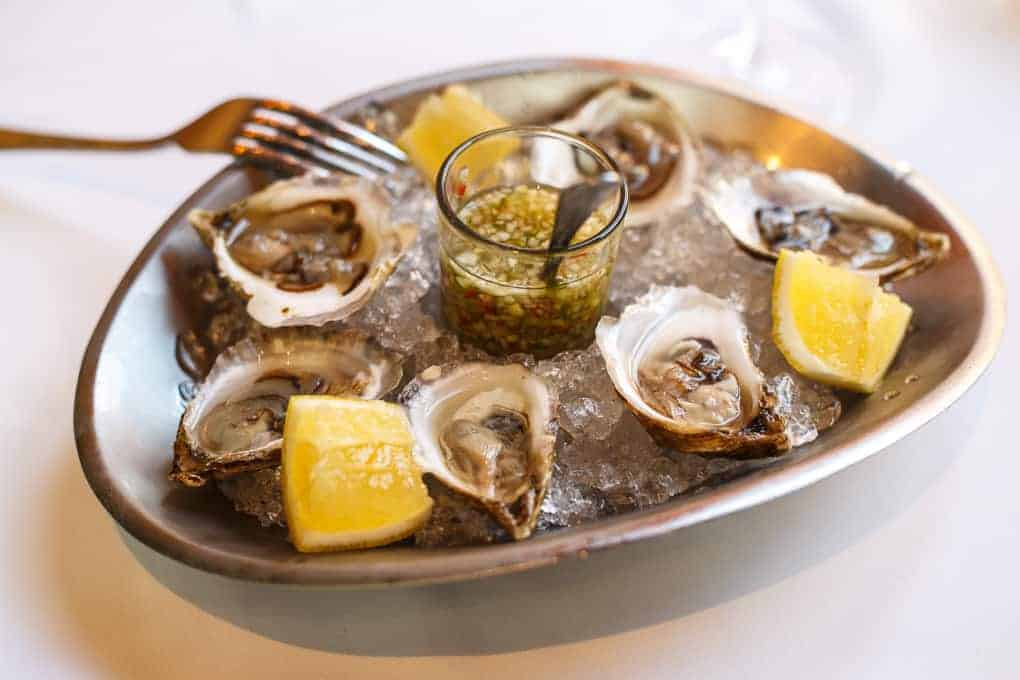 A metal plate with six raw oysters on half shells, served on crushed ice with lemon wedges and a small glass container of sauce in the center; a fork rests on the plate edge.